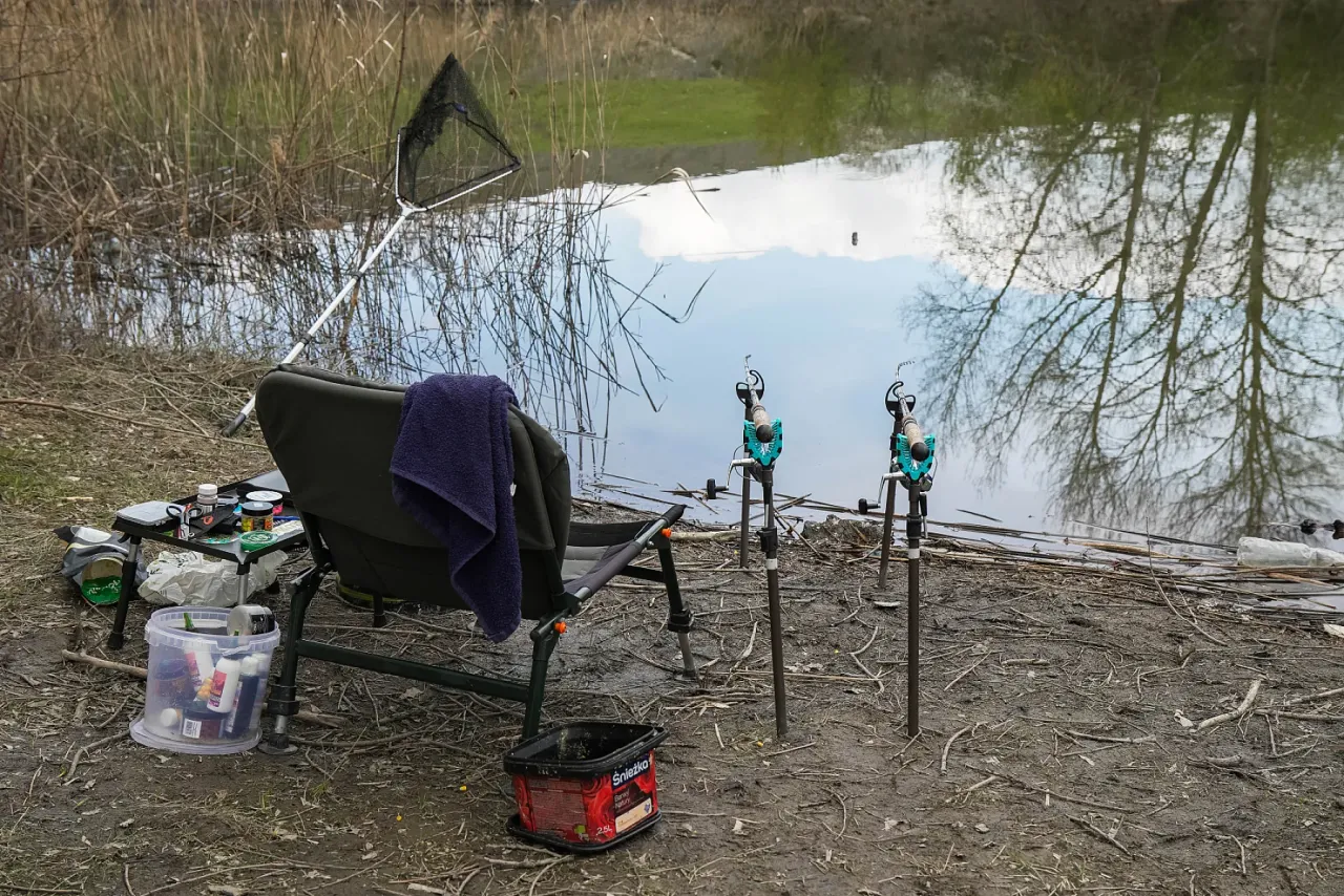 Wędka i krzesło nad wodą, gotowe do łowienia. Na stoliku obok krzesła widać pojemniki z przynętami, które mogą być najlepszą zanętą do method feeder.