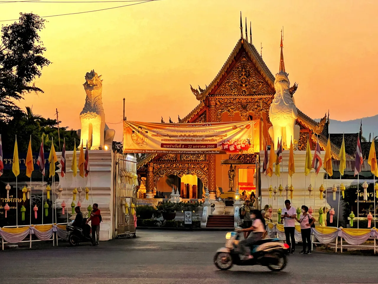 Zachód słońca nad świątynią w Chiang Mai, gdzie nocować warto w pobliżu. Złote zdobienia, lwy strażnicze i flagi tworzą magiczny widok.