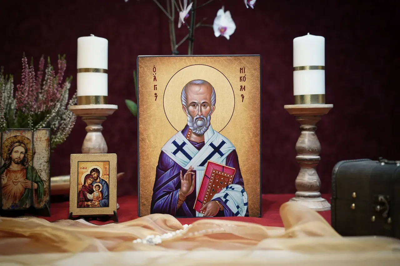 Ikona św. Mikołaja, jak z baśni, otoczona świecami i kwiatami. Obok mała ikona Świętej Rodziny i wizerunek Jezusa.
