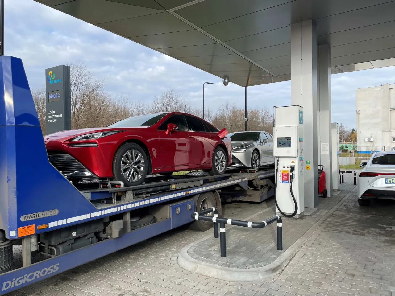 Nowe auta na wod&oacute;r czekają na tankowanie na stacji Neso. Czerwony sedan na lawecie i srebrny samoch&oacute;d obok.