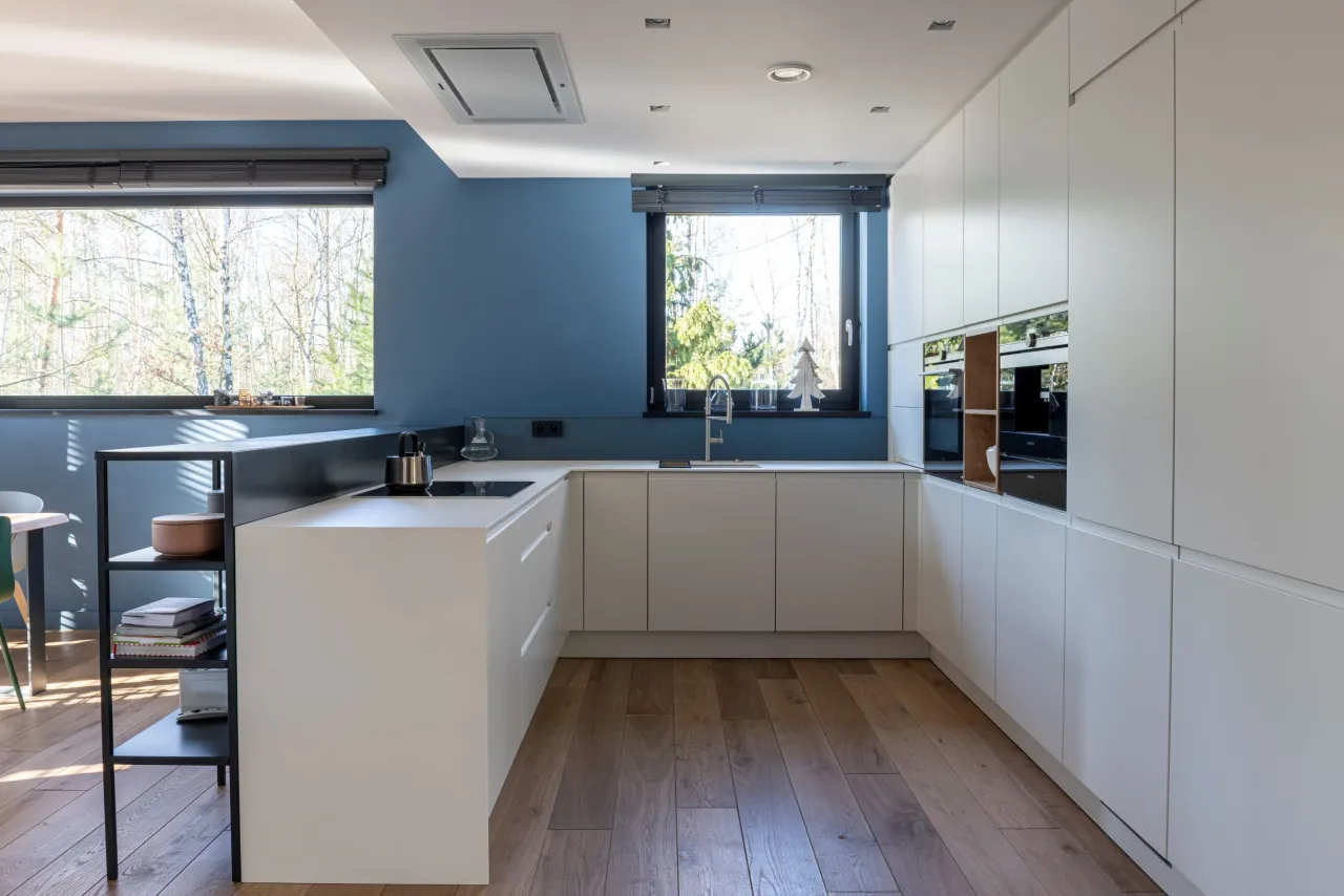 Nowoczesna kuchnia z białymi frontami i niebieską ścianą. Okna w kuchni są na wysokości blatu, zapewniając widok na drzewa.