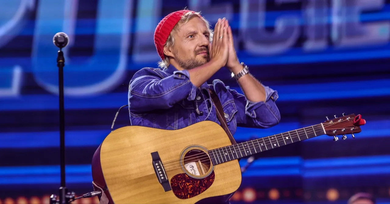 Artysta w czerwonej czapce i dżinsowej kurtce z gitarą, z dłońmi złożonymi do podziękowania. Ciekawe, ile trwa koncert Domagały?