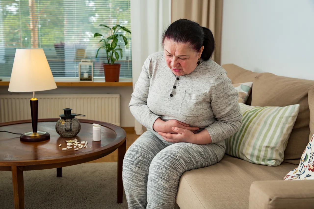 Starsza kobieta z b&oacute;lem brzucha, szukająca odpowiedzi na pytanie: jak zatrzymać biegunkę u starszej osoby. Na stoliku leki.