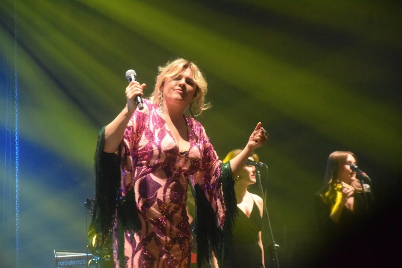 Ania Dąbrowska na scenie, w błyszczącej sukience, śpiewa do mikrofonu. Koncert trwa, a fani zastanawiają się, ile trwa koncert Ani Dąbrowskiej.