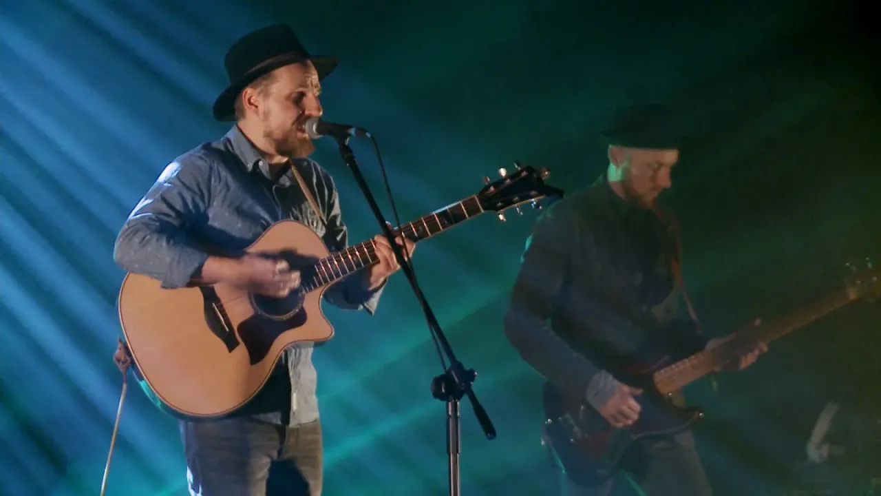 Muzyk w kapeluszu gra na gitarze akustycznej, śpiewając do mikrofonu. Obok drugi muzyk gra na gitarze basowej. Koncert Domagały trwa.