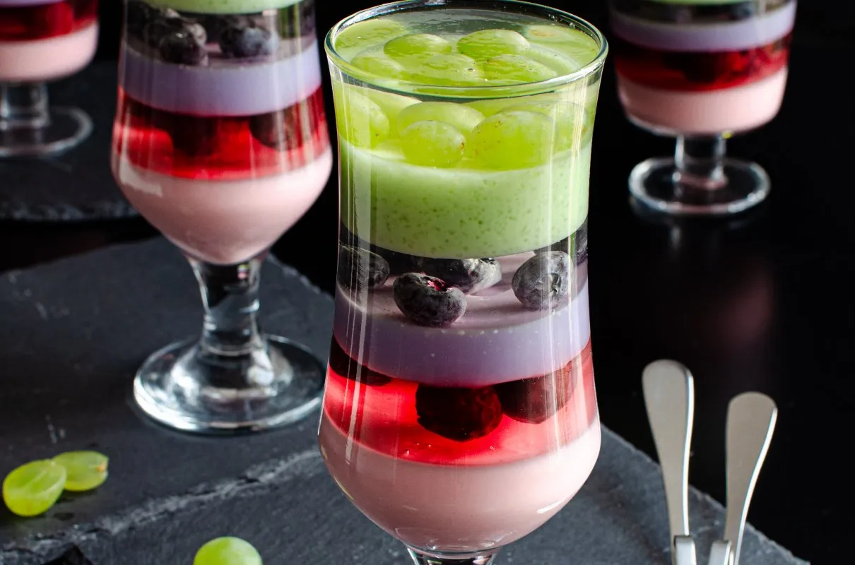 Kolorowy deser z galaretki z owocami, idealny przepis na lato. Warstwy różowej, czerwonej, fioletowej i zielonej galaretki z winogronami i borówkami.