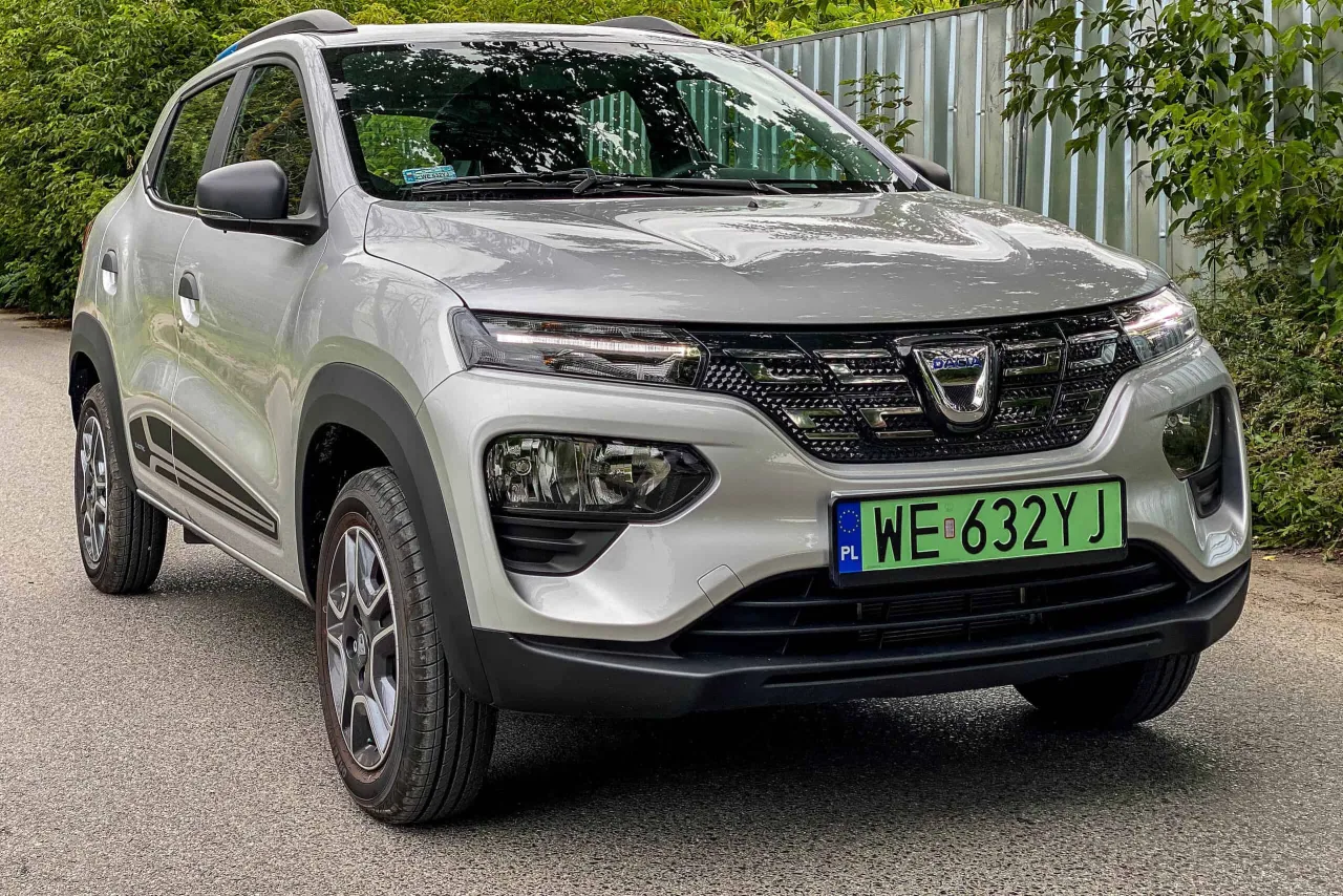 Srebrny Dacia Spring, czyli ile kosztuje najtańszy samoch&oacute;d elektryczny, zaparkowany na tle zieleni.