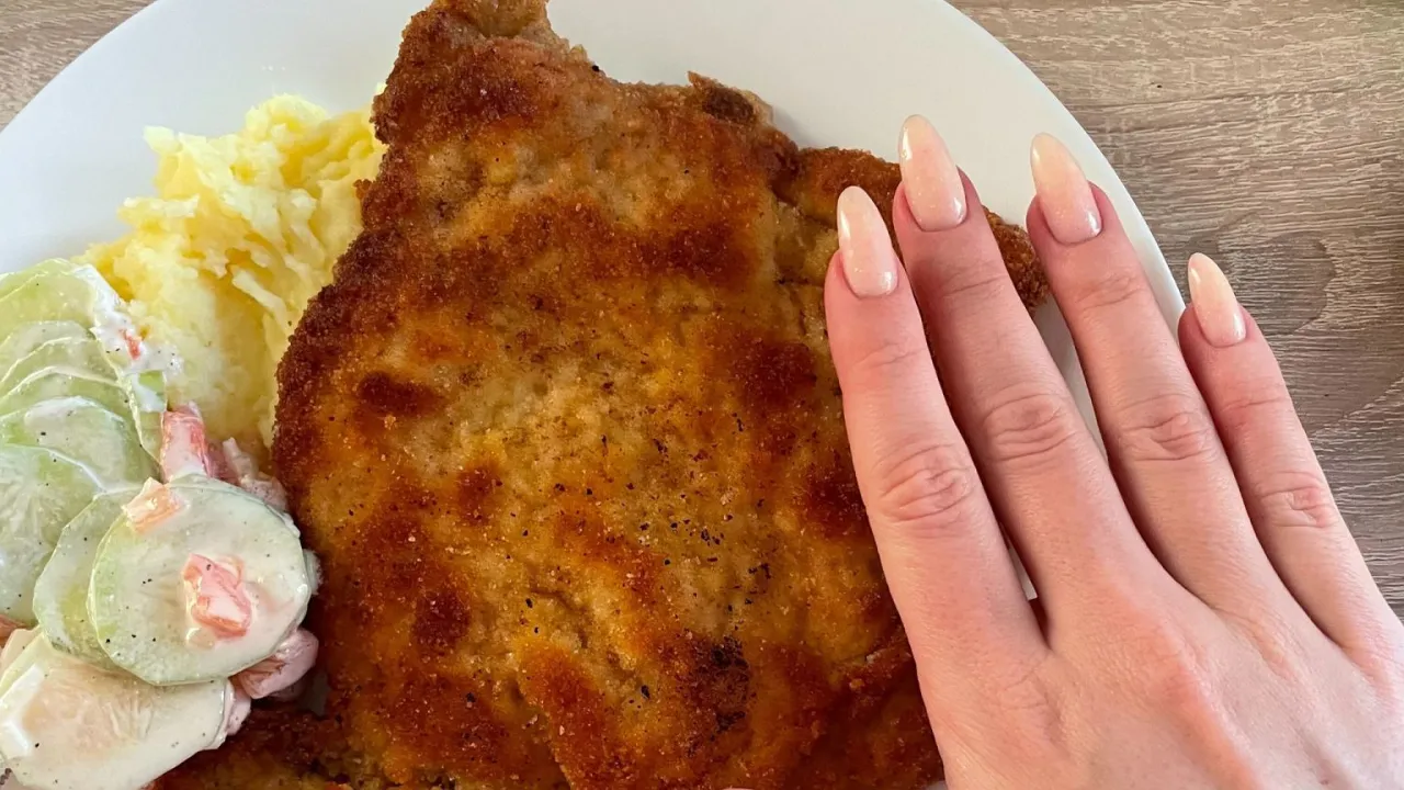 Schabowy smażony na smalcu, a na oleju roślinnym, podany z ziemniakami i mizerią. Palce z paznokciami w cielistym kolorze dotykają panierki.