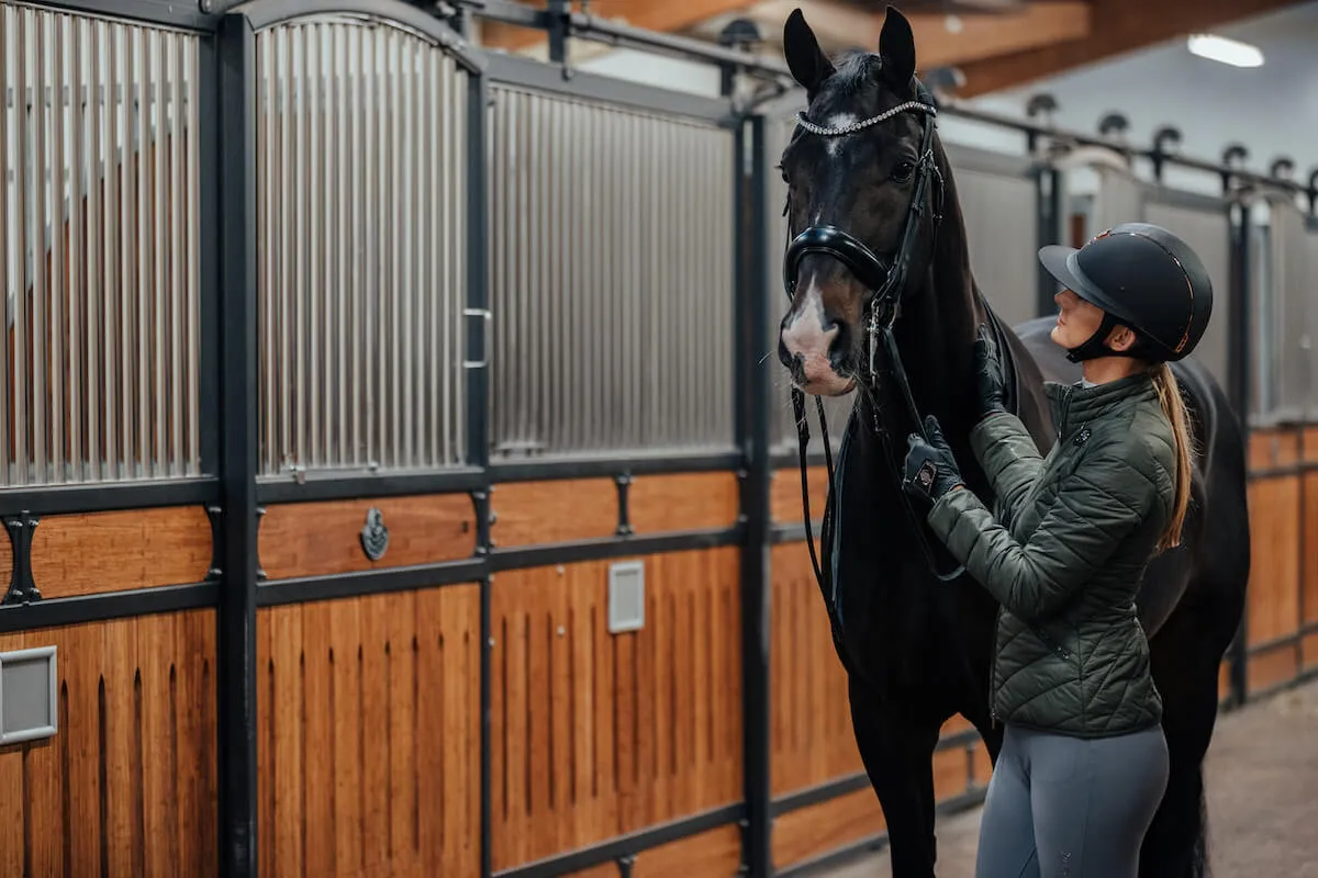 Kobieta w kasku i kurtce przygotowuje konia w stajni. Czy jazda konna jest bezpieczna? Odpowiedź zależy od przygotowania i sprzętu.