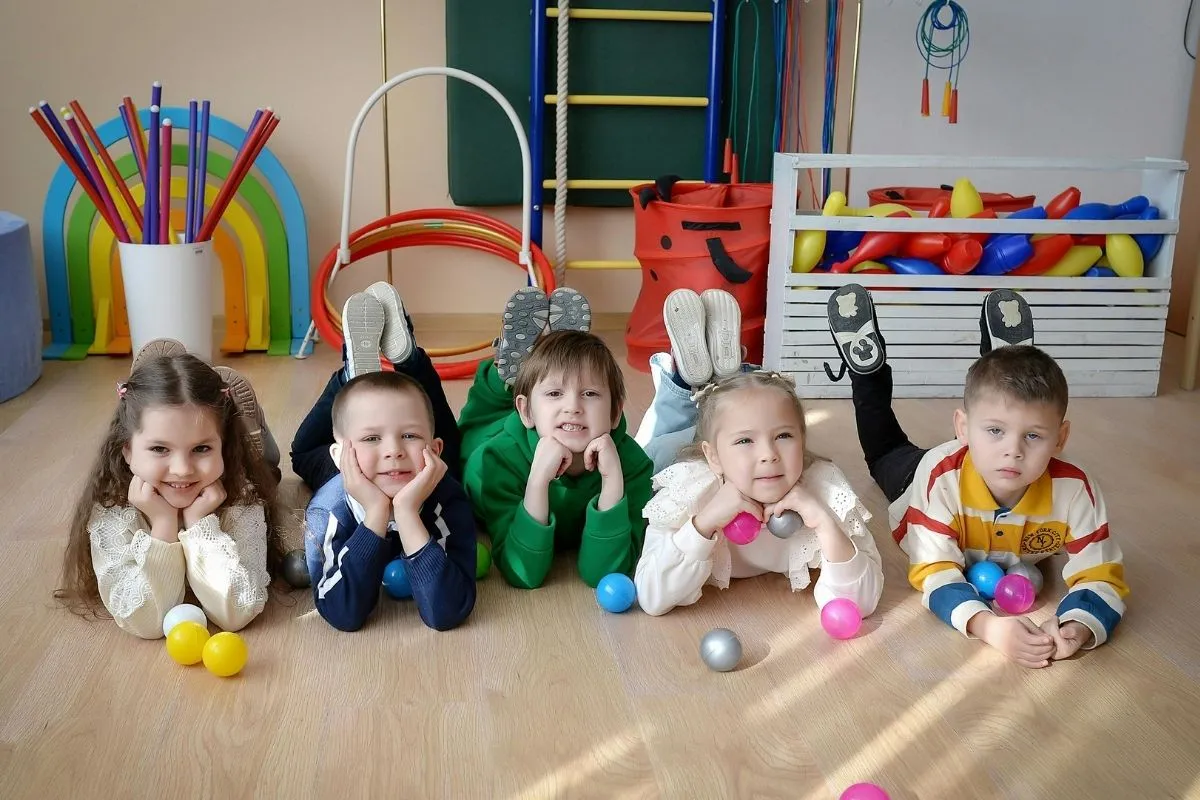 Uśmiechnięte dzieci leżą na podłodze w żłobku, bawiąc się piłeczkami. Adaptacja w żłobku przebiega pomyślnie, gdy maluchy czują się bezpiecznie i swobodnie.