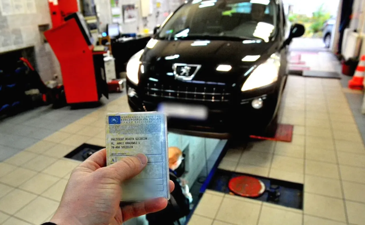 Przegląd auta w warsztacie. W ręce trzymam dowód rejestracyjny czarnego Peugeota, który czeka na diagnostykę.