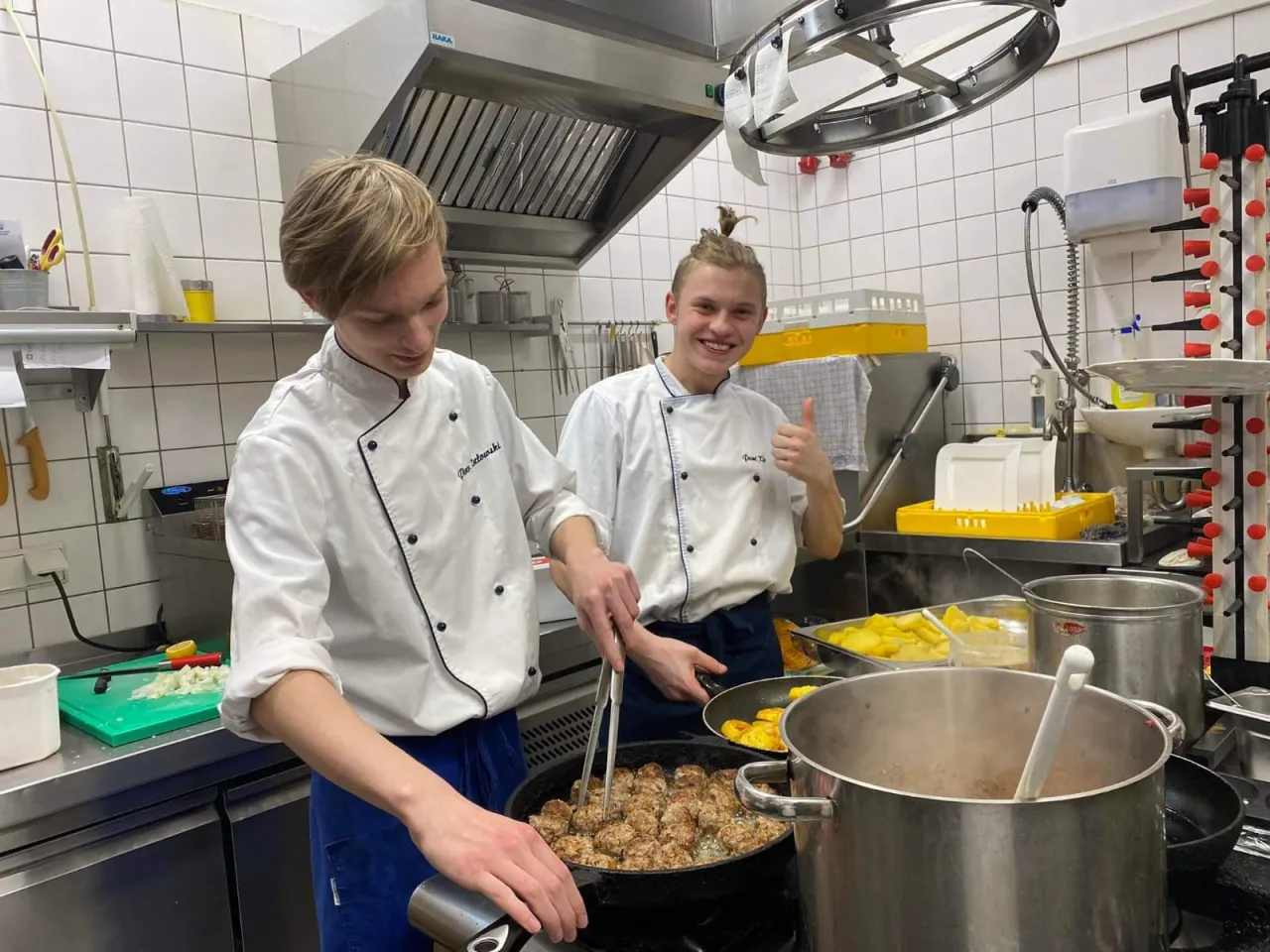 Dwóch młodych kucharzy w kuchni. Jeden miesza klopsiki na patelni, drugi pokazuje kciuk w górę. Kod zawodu kucharz to ich pasja.