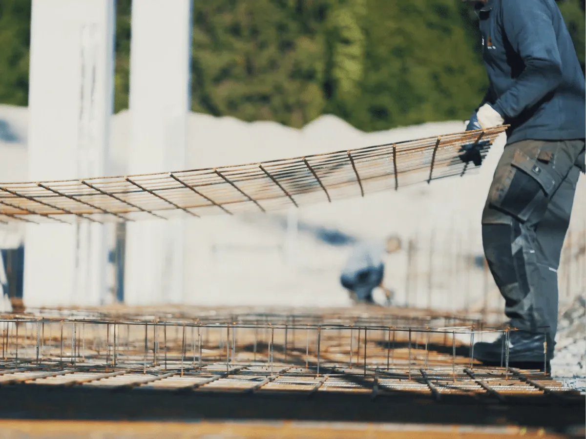 Budowlaniec układa siatkę zbrojeniową. W tle widać inne elementy konstrukcyjne i pracujących ludzi. Dystanse do zbrojenia zapewniają odpowiednią grubość otuliny betonowej.