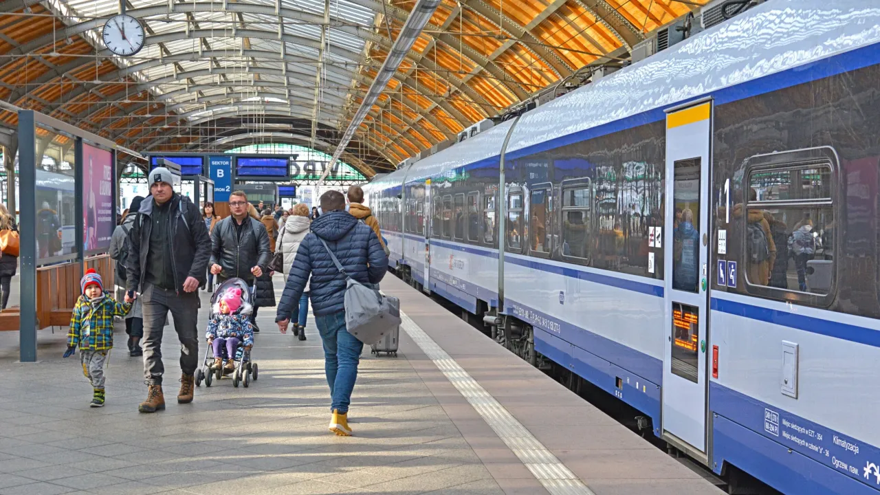 Rodzina z dziećmi na peronie, czekająca na pociąg Intercity. W tle widoczny zegar i tablice informacyjne.
