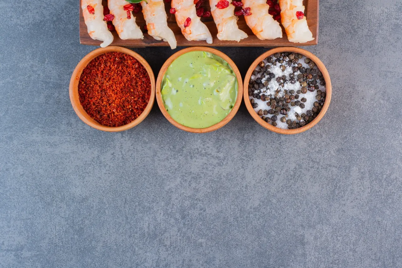 Trzy miseczki z przyprawami do ramen: chili, zielony sos i pieprz z solą. Obok krewetki.