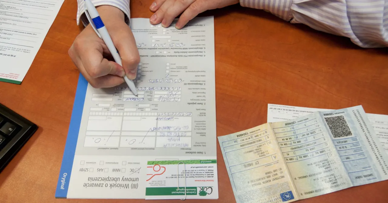 Ręka wypełnia wniosek o ubezpieczenie, szukając sposob&oacute;w na tanie OC. Obok dokumenty pojazdu.