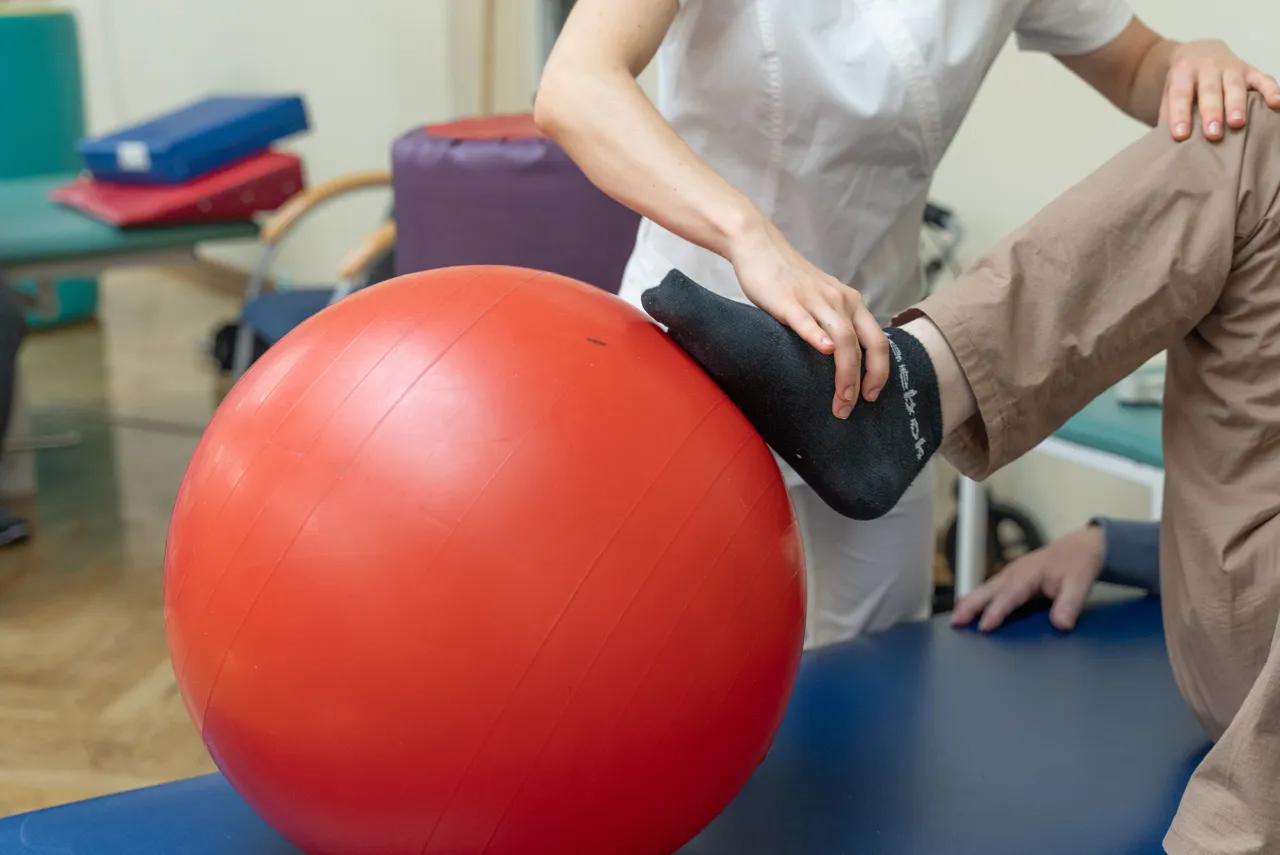 Fizjoterapeuta pomaga pacjentowi w ćwiczeniach na dużej, czerwonej piłce. Rehabilitacja ud na czym polega? Na poprawie siły i ruchomości.