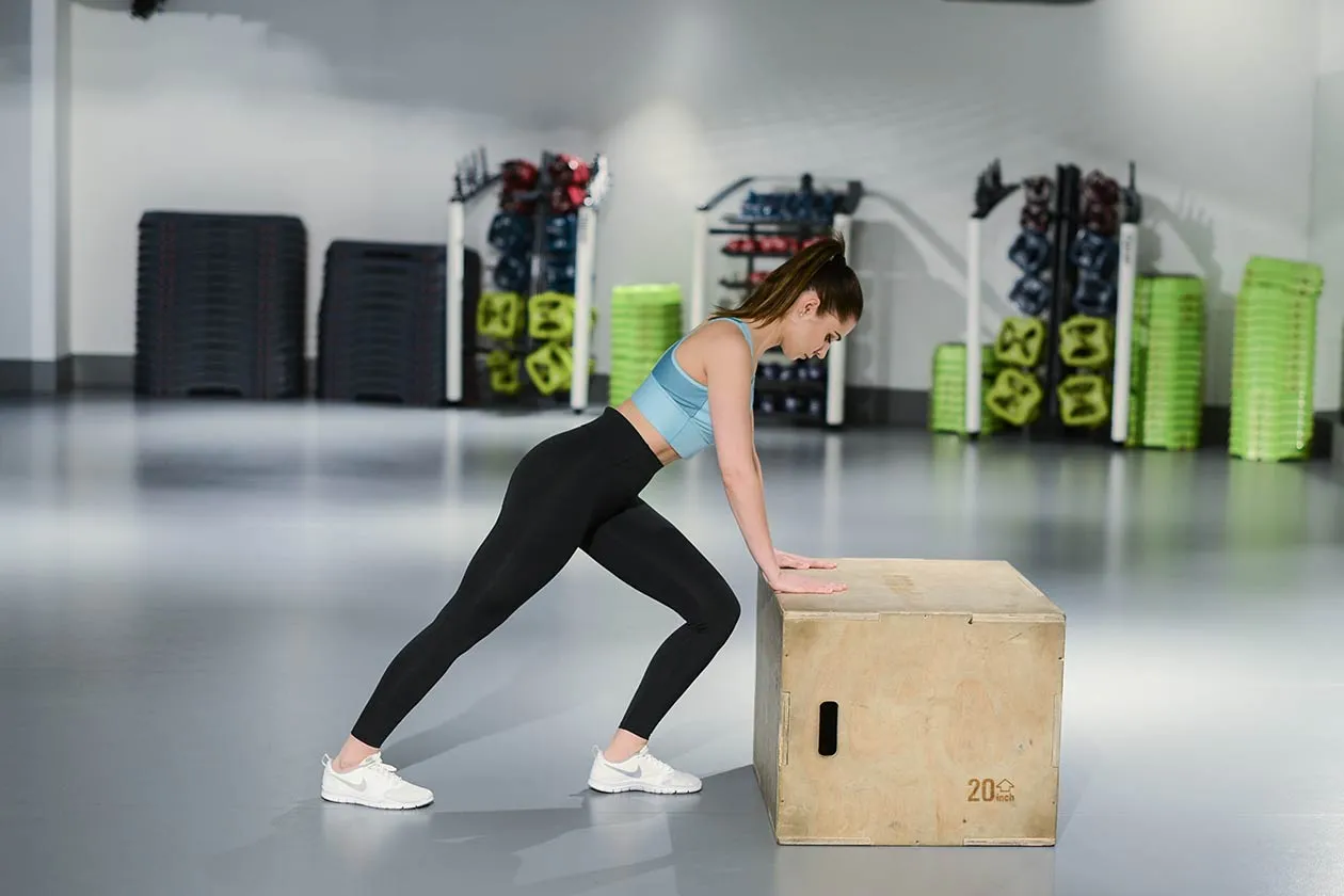 Kobieta ćwiczy, wykonując ćwiczenie na skrzyni, które pomaga jak wysmuklić łydki. W tle sprzęt do ćwiczeń.