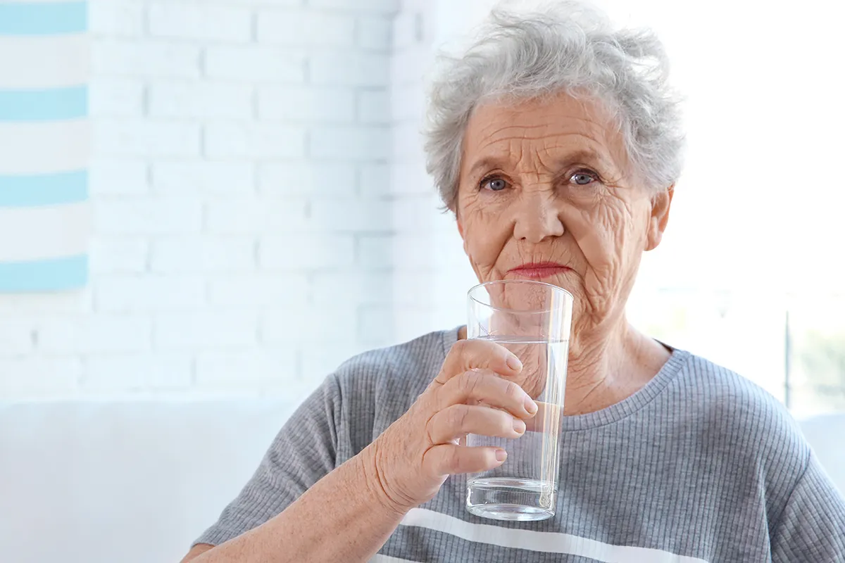 Starsza pani pije wodę, co jest dobrym sposobem na zapobieganie odwodnieniu u os&oacute;b starszych.