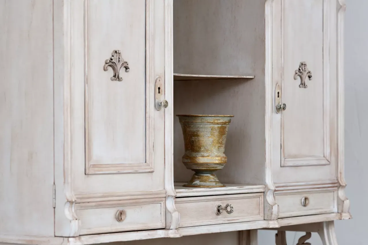 Odkryj, jak odnowić stare meble, by nadać im nowy blask. Biała komoda z ozdobnymi uchwytami i złotą wazą w środku.