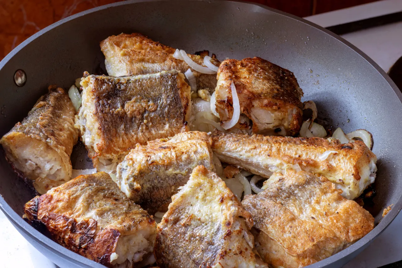 Kawałki ryby smażą się na patelni z cebulą. Złociste, chrupiące kawałki sugerują, że czas smażenia ryby na patelni jest optymalny.