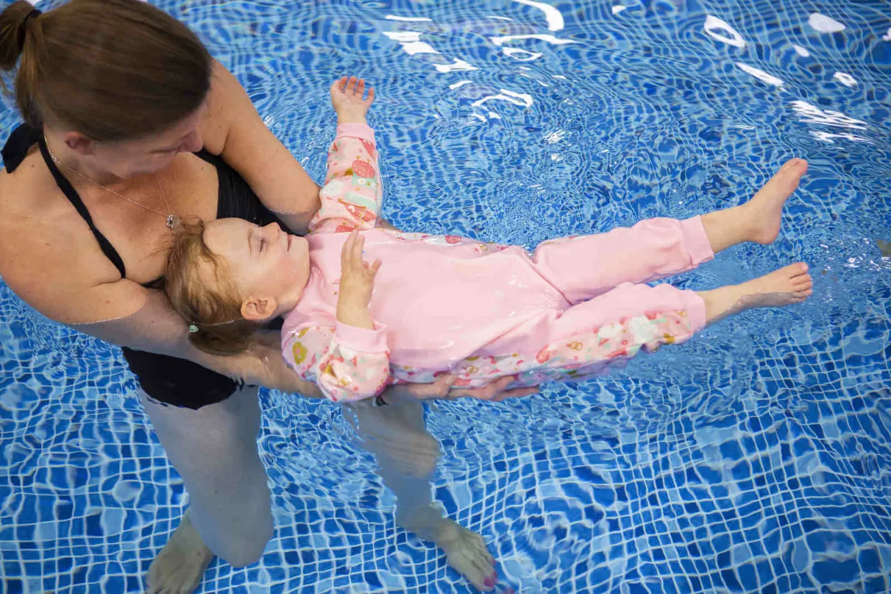 Dziecko w r&oacute;żowym kombinezonie do pływania dla dzieci unosi się na wodzie, trzymane przez instruktorkę.