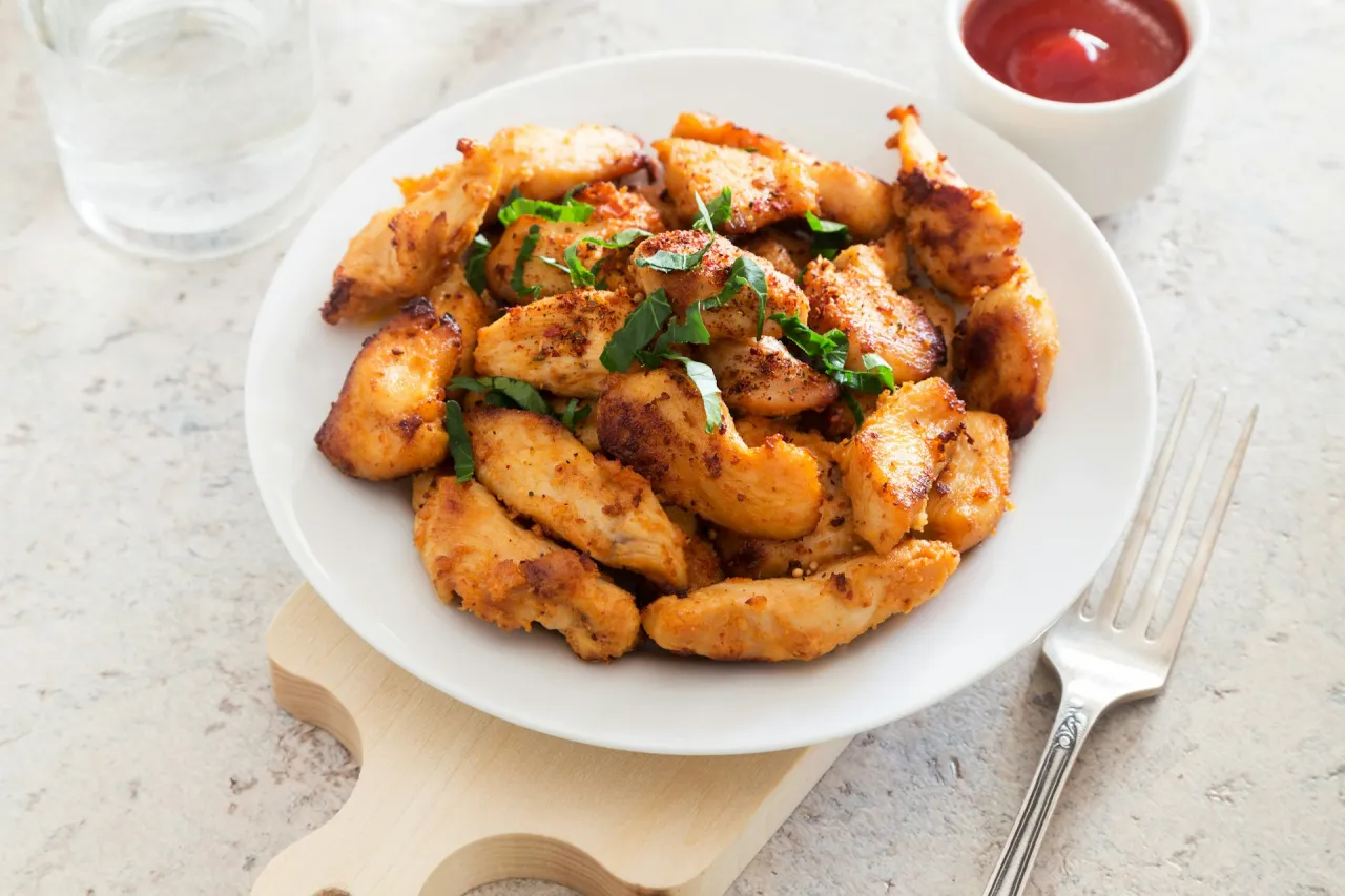 Talerz pełen chrupiących kawałk&oacute;w kurczaka, posypanych ziołami. Idealny obiad z dużą ilością białka, obok sos i szklanka wody.