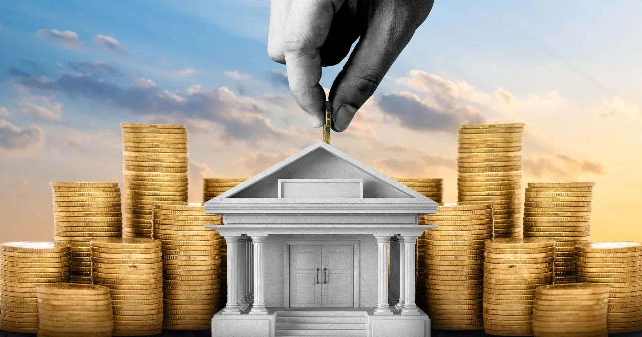 Dłoń wrzuca monetę do budynku banku otoczonego stosami złota. To symbol bezpieczeństwa finansowego, czyli co to jest fundusz gwarancyjny.
