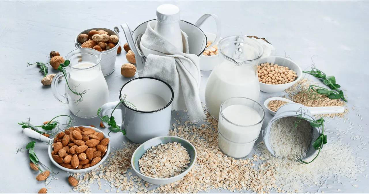 Wyb&oacute;r między mlekiem sojowym czy owsianym? Na stole mleko migdałowe, ryżowe, z orzech&oacute;w i soi, obok płatki owsiane i migdały.