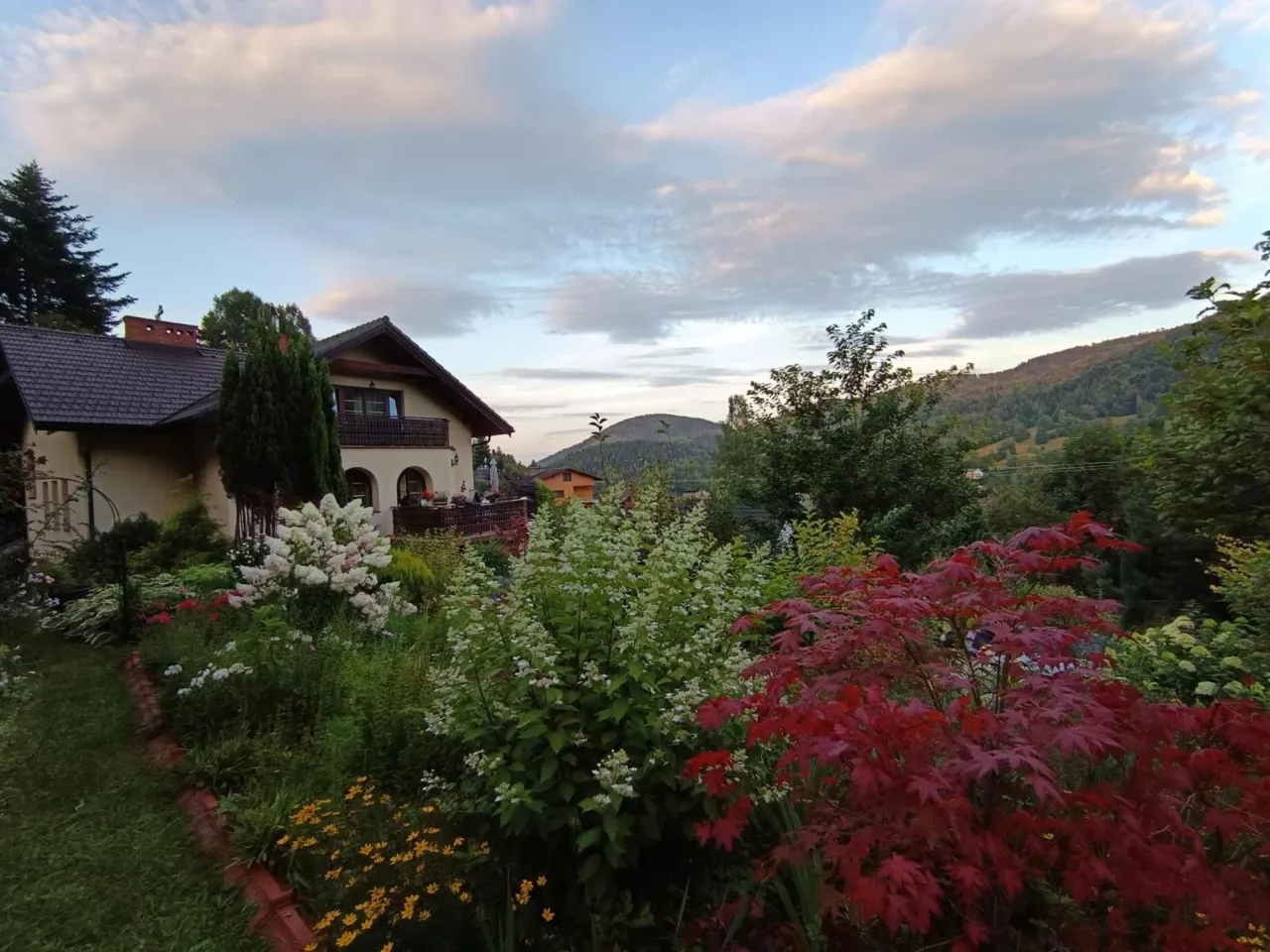 Willa Wierchy w Szczyrku otoczona kwitnącym ogrodem. W tle g&oacute;ry i niebo z chmurami.