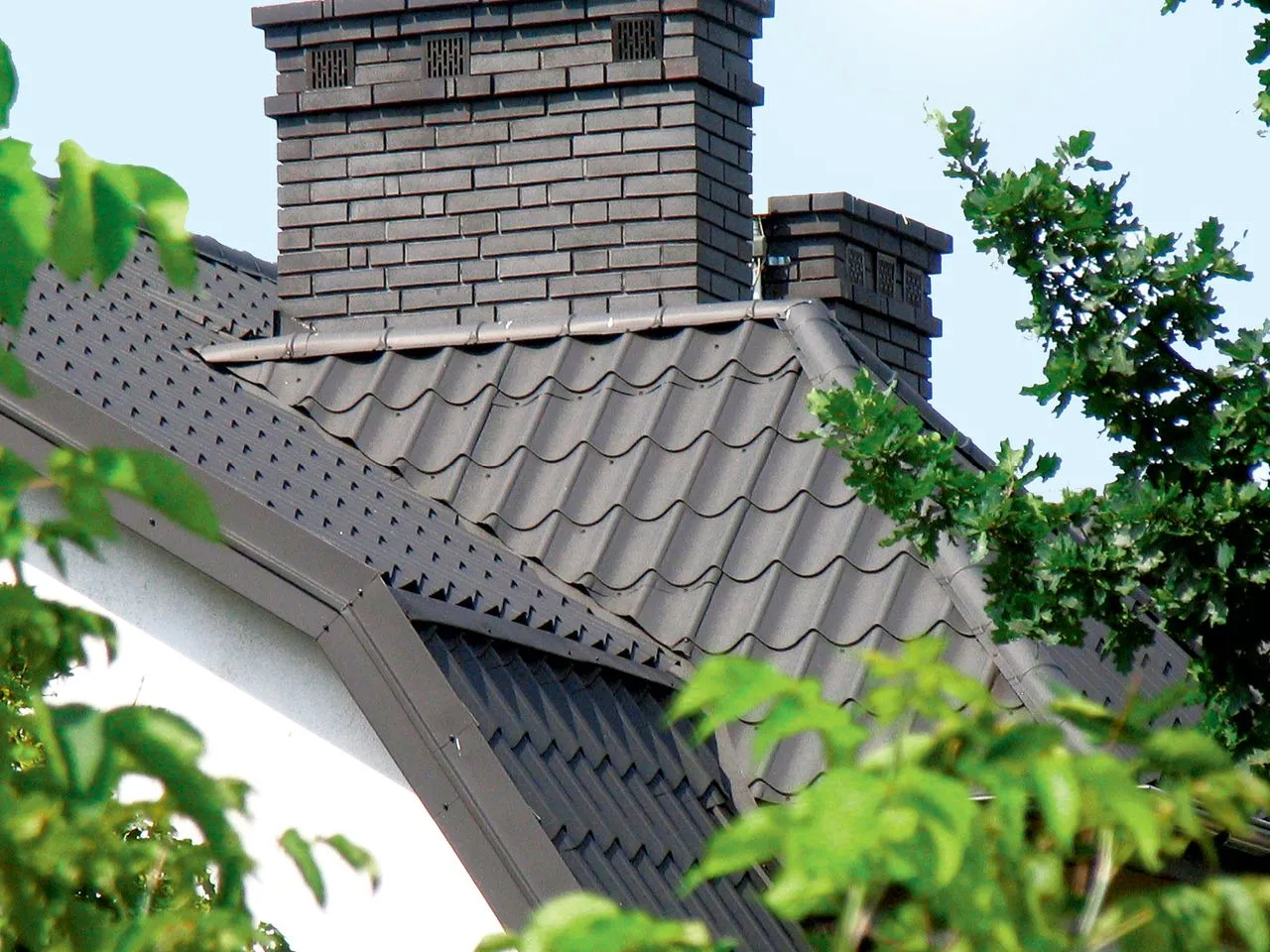 Dwa ceglane kominy do co, dach pokryty ciemną dach&oacute;wką, zielone liście drzew.