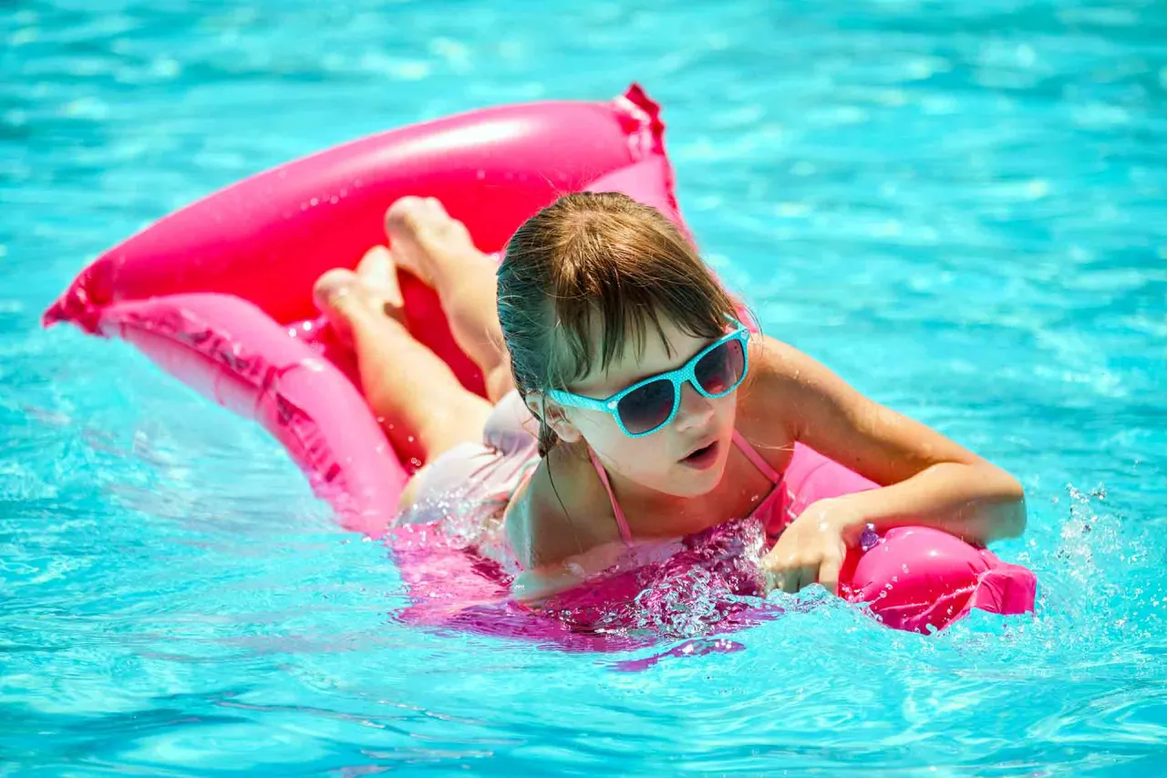 Dziewczynka w okularach przeciwsłonecznych pluska się w basenie na r&oacute;żowym materacu do pływania.