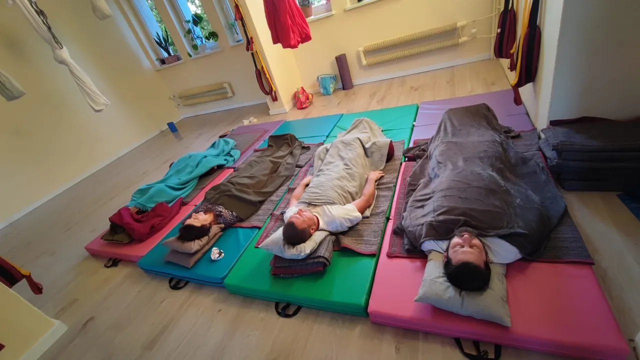 Ludzie leżą na matach, przykryci kocami, podczas relaksujących ćwiczeń jogi nidry.