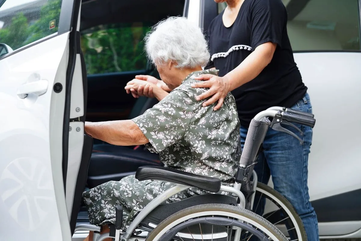 Opiekunka pomaga starszej kobiecie wstać z w&oacute;zka inwalidzkiego do samochodu. Czy opieka nad starszą osobą wlicza się do lat pracy?