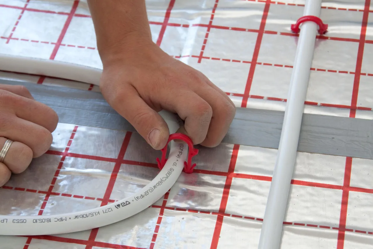 Ręce układają białą rurę na folii izolacyjnej, mocując ją czerwonym klipsem. To kluczowy etap, jak podłączyć ogrzewanie podłogowe.
