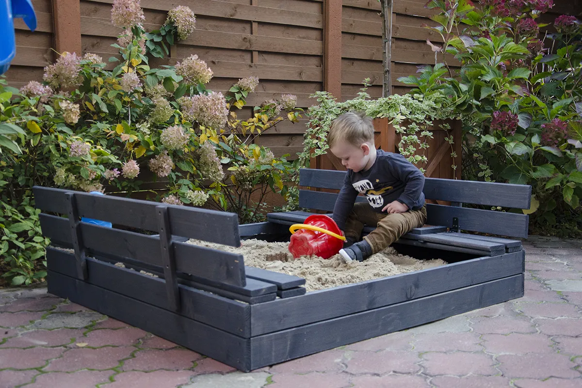 Chłopiec bawi się w piaskownicy z ławkami, która wygląda jakby właśnie skończono ją budować. To świetny pomysł, jak zrobić piaskownicę dla dzieci.