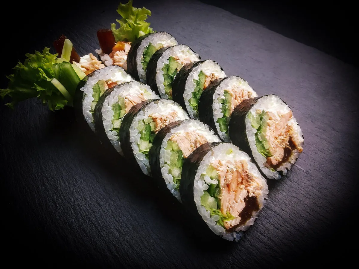 Rząd futomaków z pieczonym łososiem, chrupiącym ogórkiem i sałatą, podane na czarnej płycie.