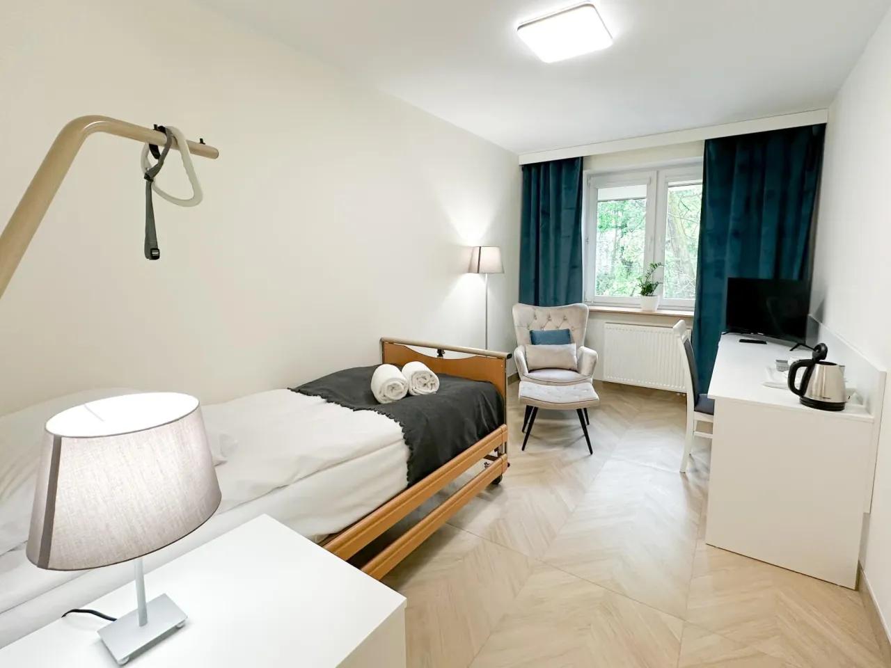 Pokój rehabilitacyjny z łóżkiem, fotelem i biurkiem. Idealne miejsce, gdzie najlepsza rehabilitacja kardiologiczna pomoże wrócić do zdrowia.