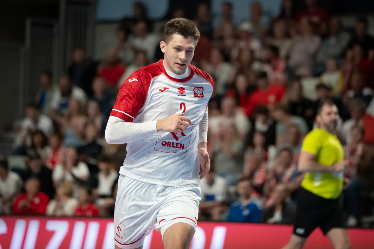 Piłkarz ręczny w białej koszulce z orłem na piersi biegnie po boisku. Kibice na trybunach czekają na kolejny mecz polski w piłce ręcznej.