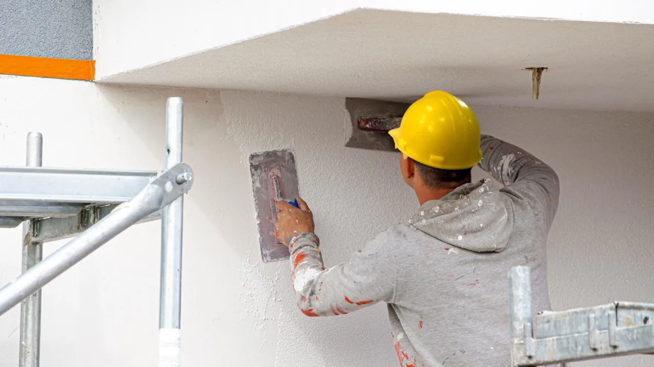 Pracownik w żółtym kasku nakłada tynk gipsowy na ścianę. Zastanawiasz się, ile za tynki gipsowe?