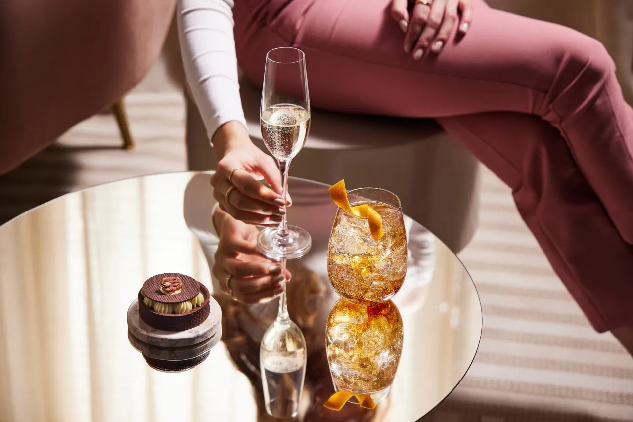 Elegancki prosecco drink i deser na lustrzanym stoliku. Kobieta w różowych spodniach sięga po kieliszek musującego napoju.