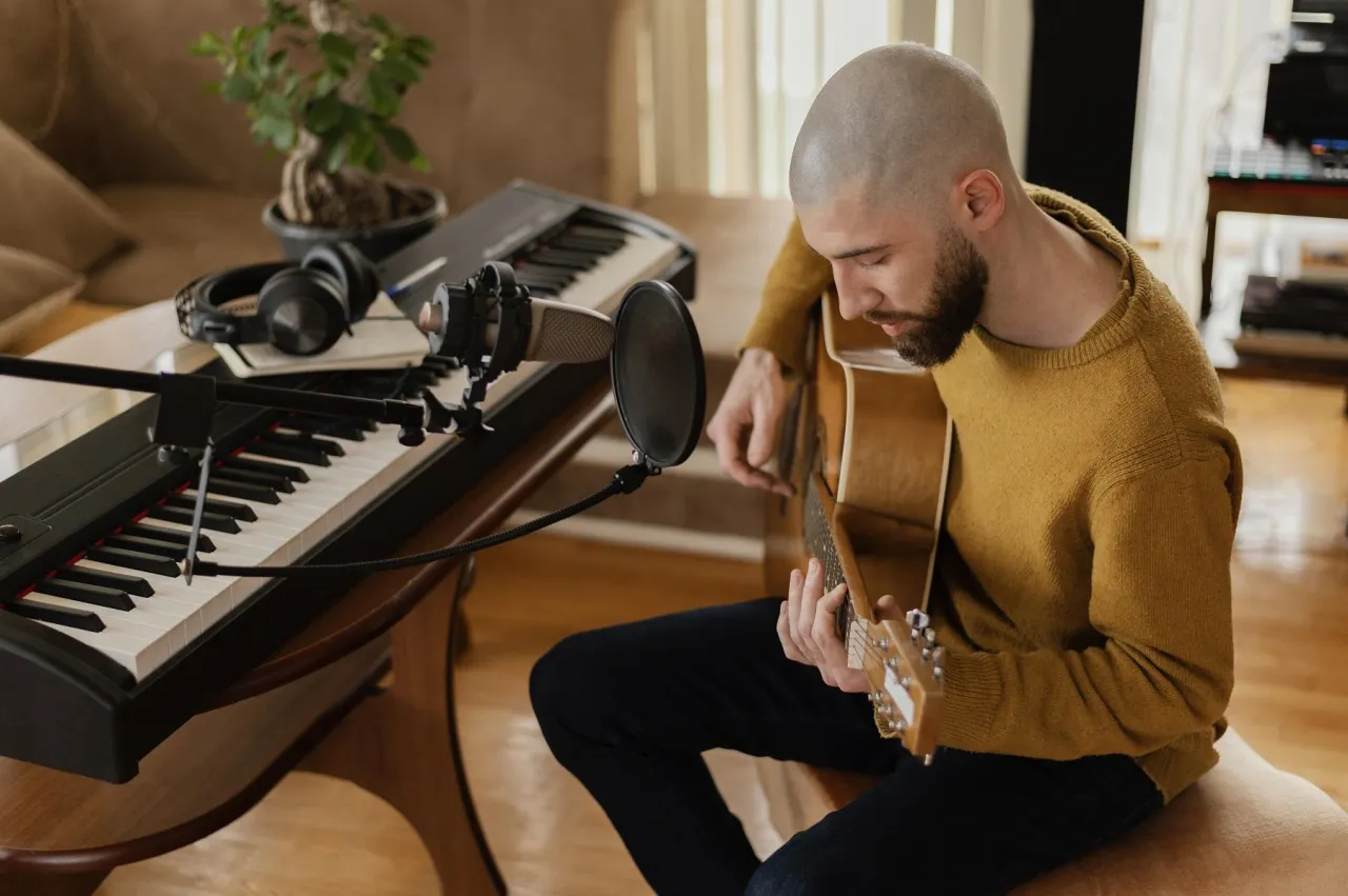 Mężczyzna gra na gitarze przy pianinie i mikrofonie. Tworzy muzykę, która pomoże mu sprzedać tekst piosenki.
