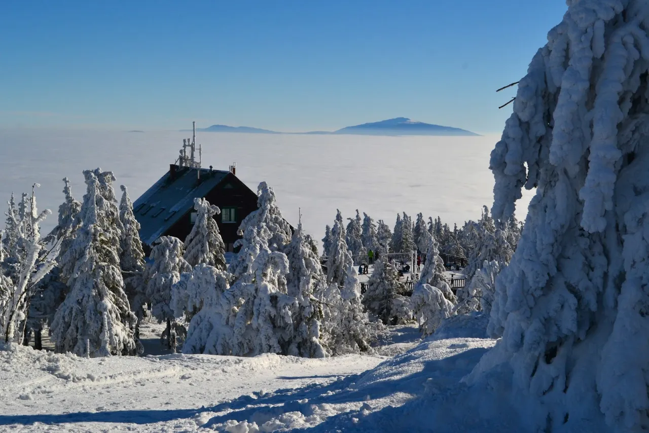Zimowy krajobraz w Istenie. Śnieżne drzewa otaczają schronisko, a w oddali widać morze chmur i zarys g&oacute;r.