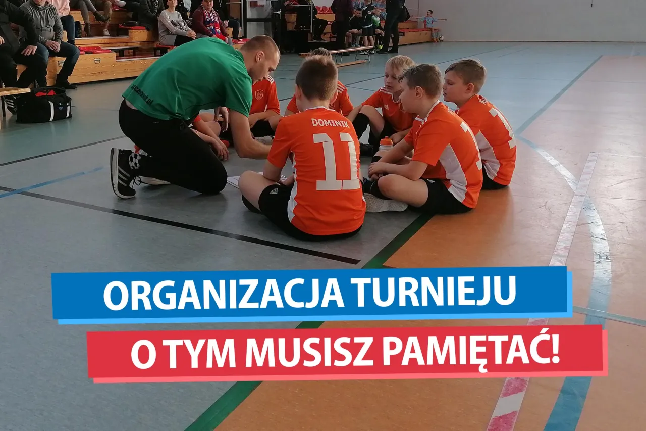 Trener w zielonej koszulce tłumaczy dzieciom, jak zorganizować turniej piłki nożnej. Wszyscy siedzą na podłodze sali gimnastycznej.