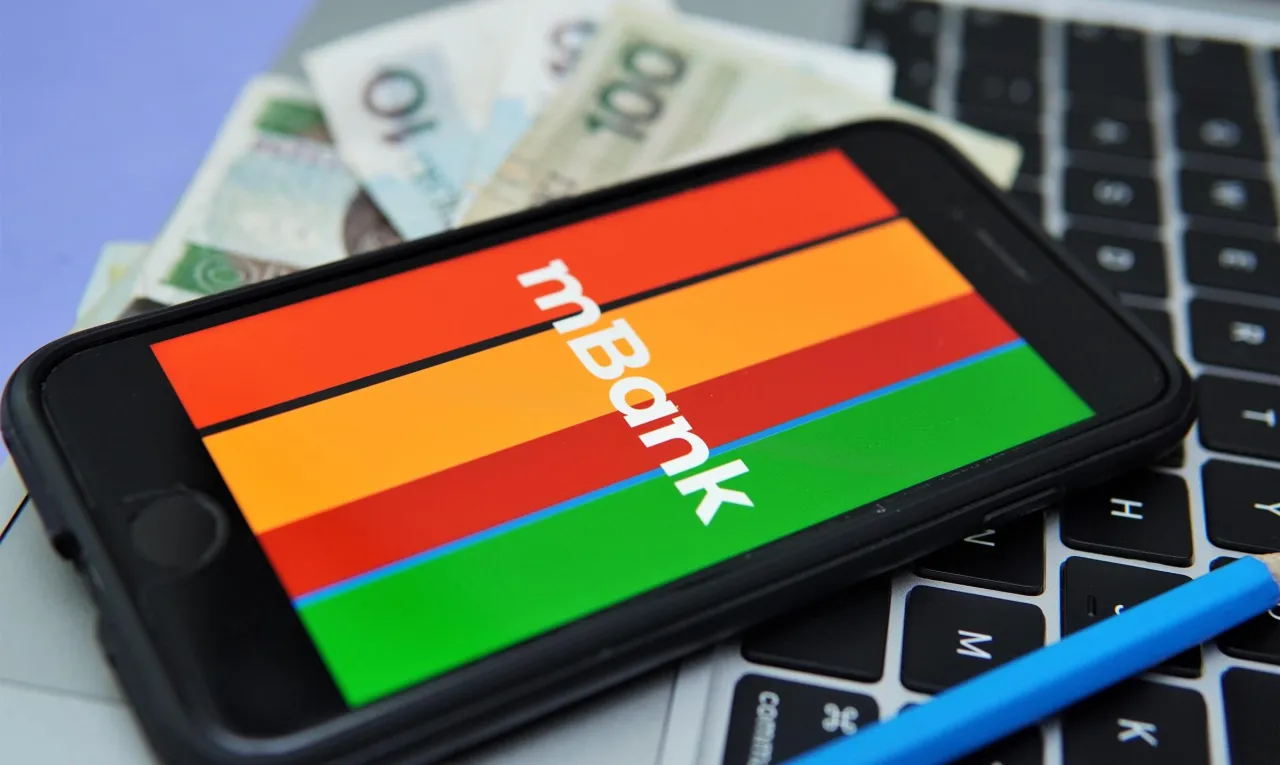Smartfon z logo mBank na klawiaturze, obok polskie banknoty. Dowiedz się, jak anulować przelew mBank.