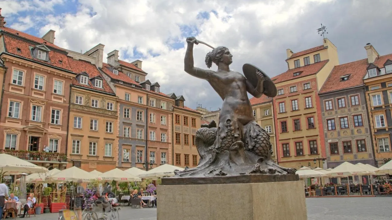 Pomnik Syrenki Warszawskiej, bohaterki legendy o syrence, stoi na tle kolorowych kamienic.