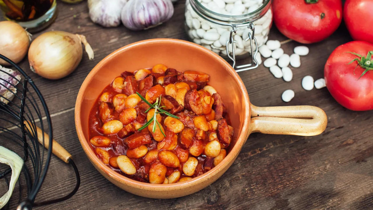 Fasola po bretońsku z kiełbasą w glinianej misce. Jakie przyprawy do fasoli po bretońsku? W tle cebula, czosnek, pomidory i słoik fasoli.
