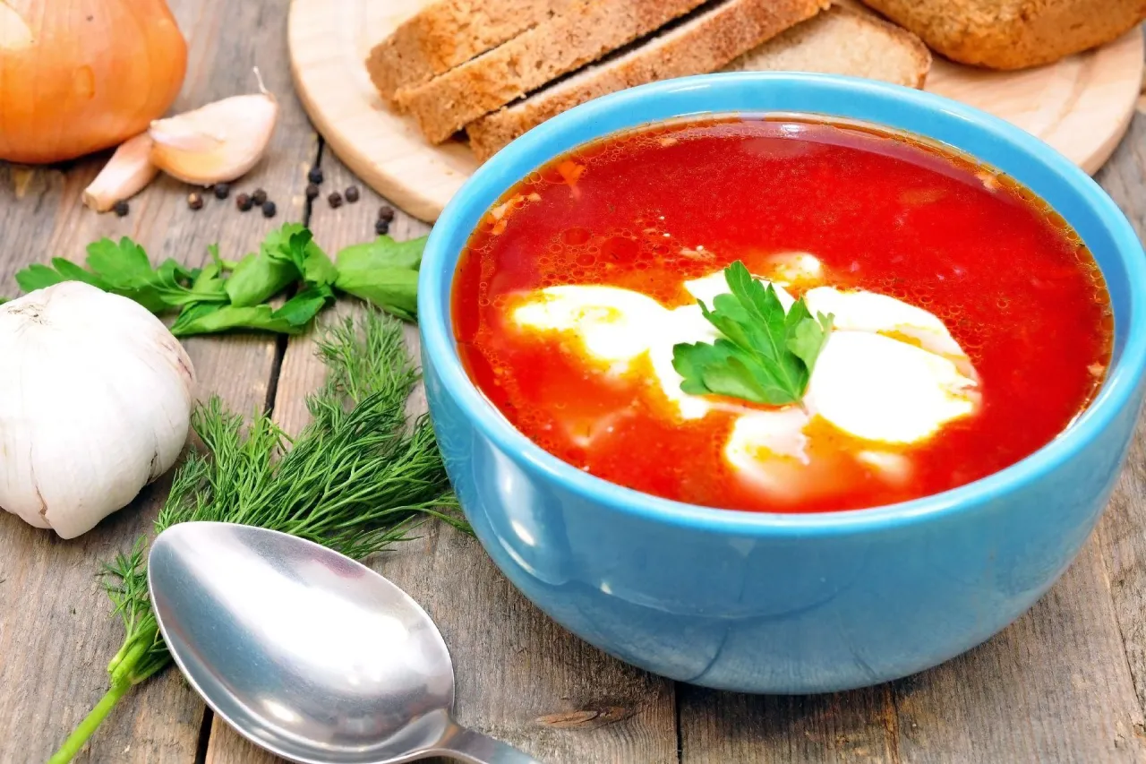 Apetyczny barszcz z kleksem jogurtu naturalnego, który pokazuje, czy można zabielić zupę jogurtem naturalnym. Obok pieczywo, czosnek i koperek.