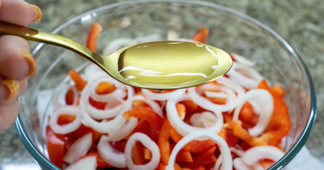 Złota łyżka z płynem, prawdopodobnie olejem, nad miską z pokrojonymi warzywami. Sterole roślinne co to? To składnik zdrowej diety.