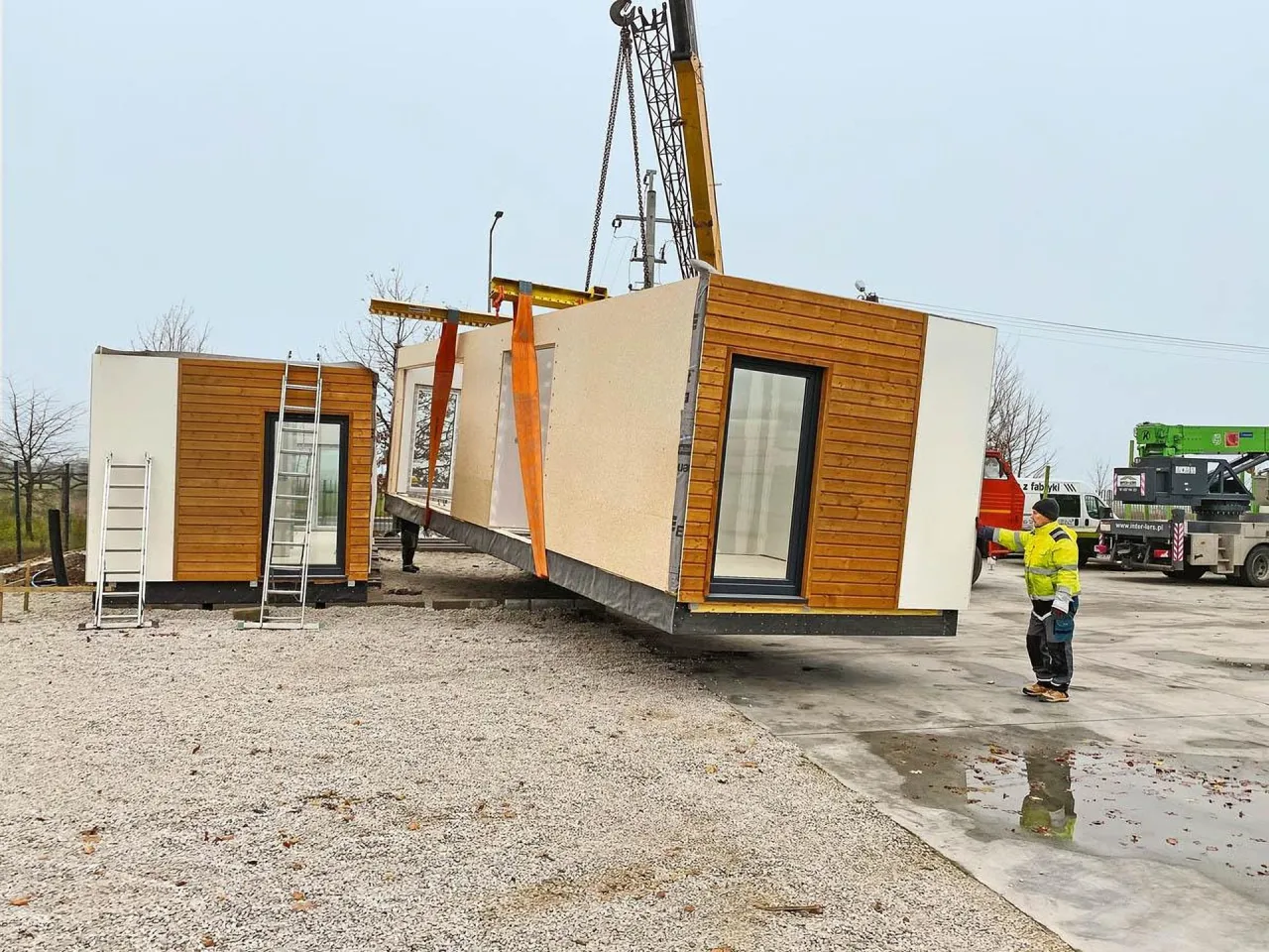 Montaż domu modułowego. Dźwig podnosi jeden z modułów, pracownik nadzoruje. To pokazuje, jak szybko powstaje dom modułowy.